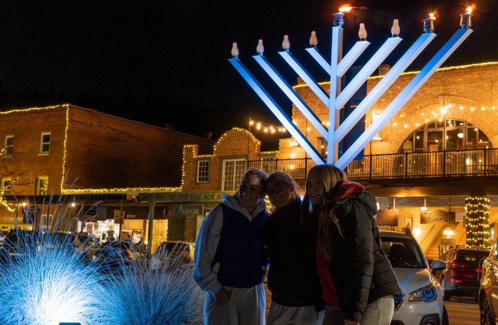 Three people stand near a large, lit menorah at night in a festive town setting.