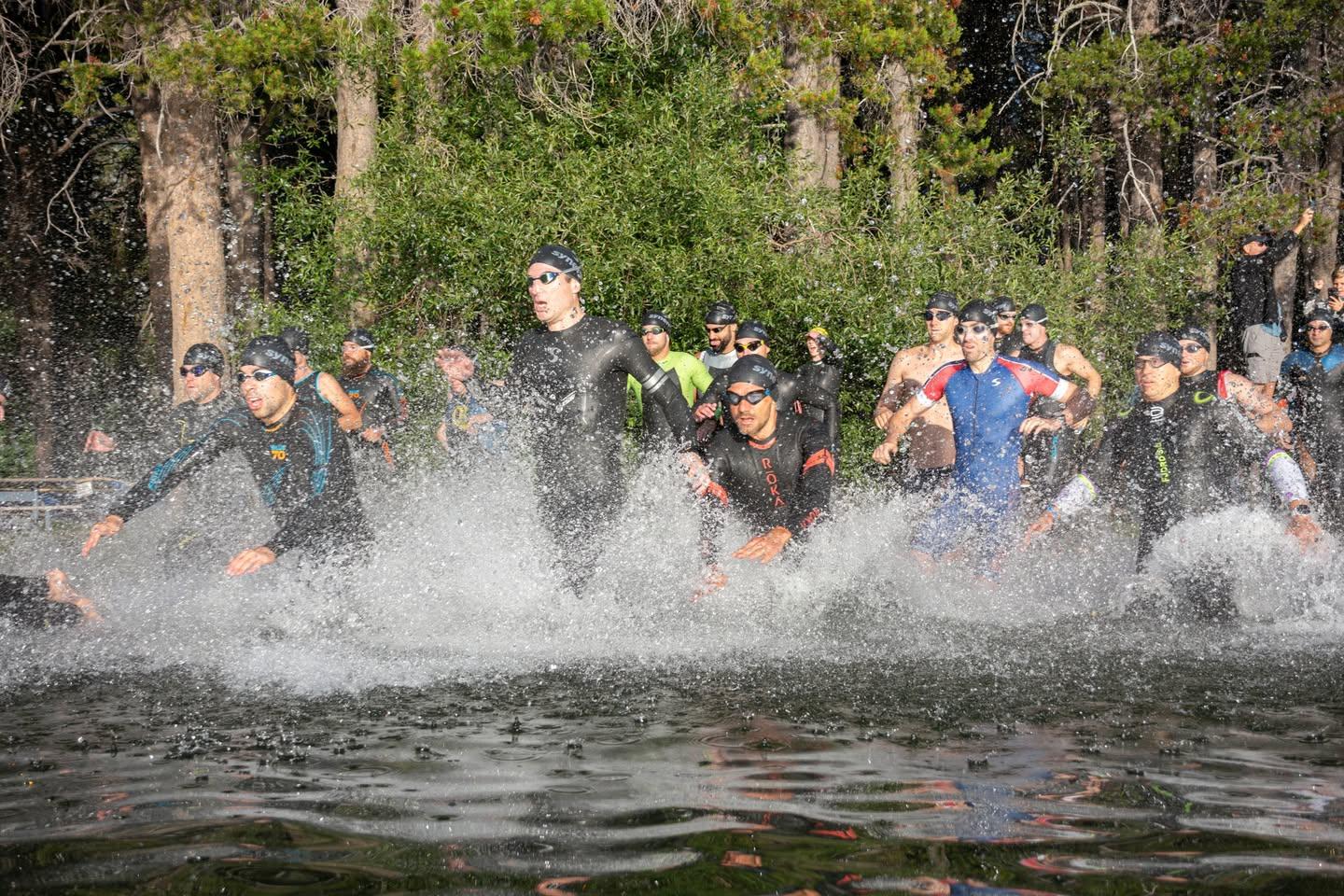 Athletes in wetsuits run into a lake, splashing water, surrounded by trees.