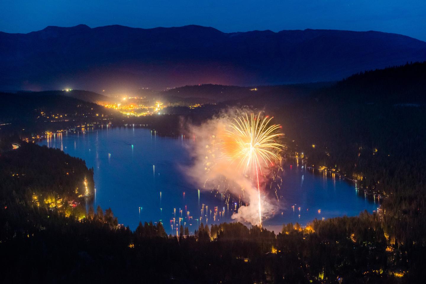 Fireworks over a lake at night, surrounded by lit-up trees and distant mountains.