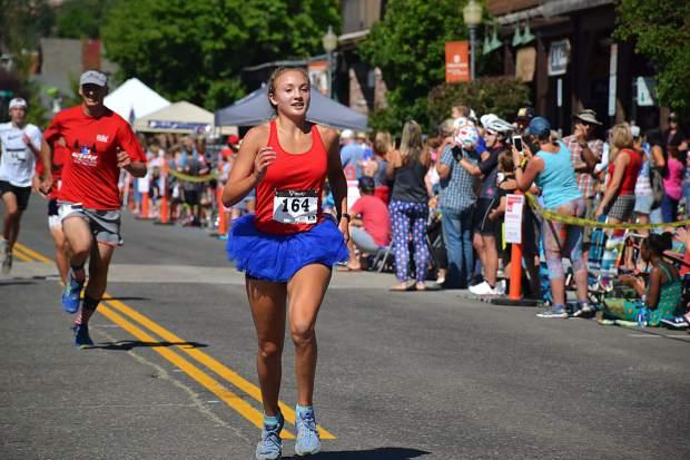 Runner in a blue tutu crossing the finish line in a road race, crowd cheering.