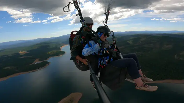 Paragliders soar above a scenic landscape, with a river and forest below.