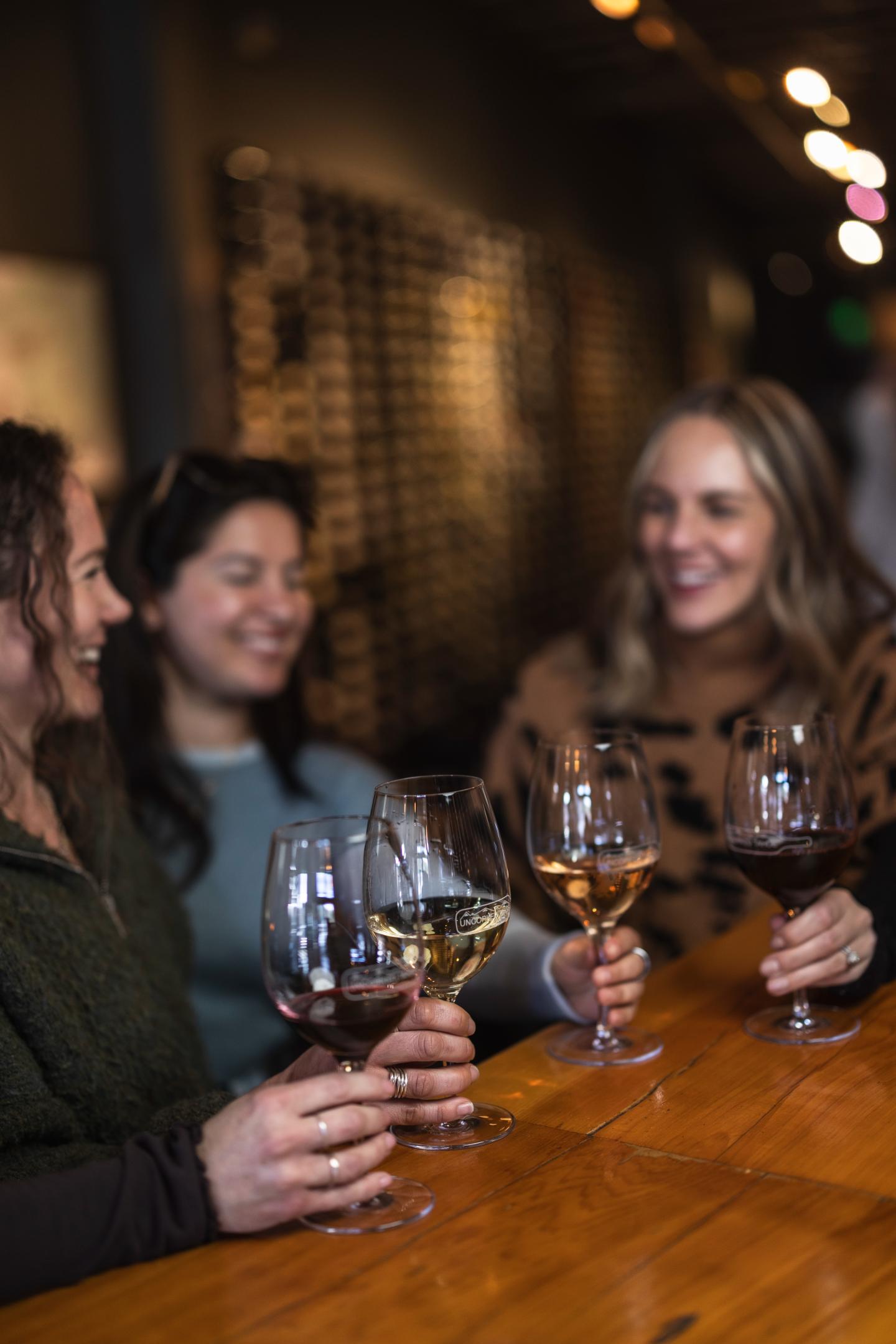 Three women smiling and clinking wine glasses at a wooden table.