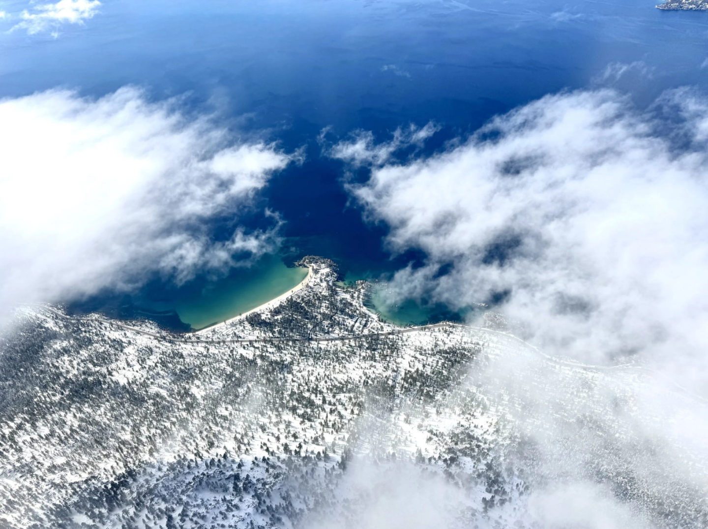 Aerial view of a snowy landscape by a deep blue lake, partially covered by clouds.