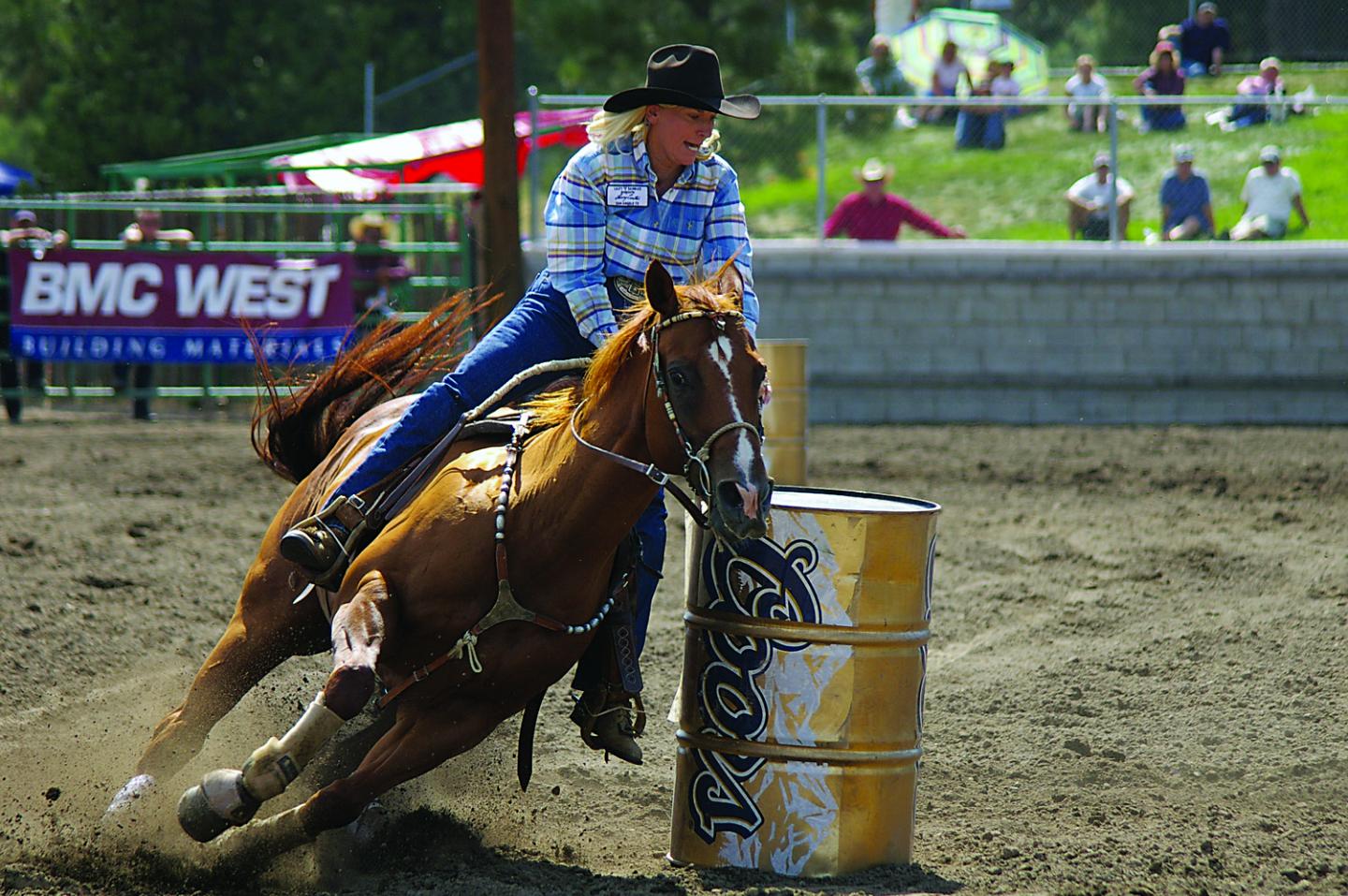 Rodeo rider in plaid shirt rounding a barrel on a horse mid-race.
