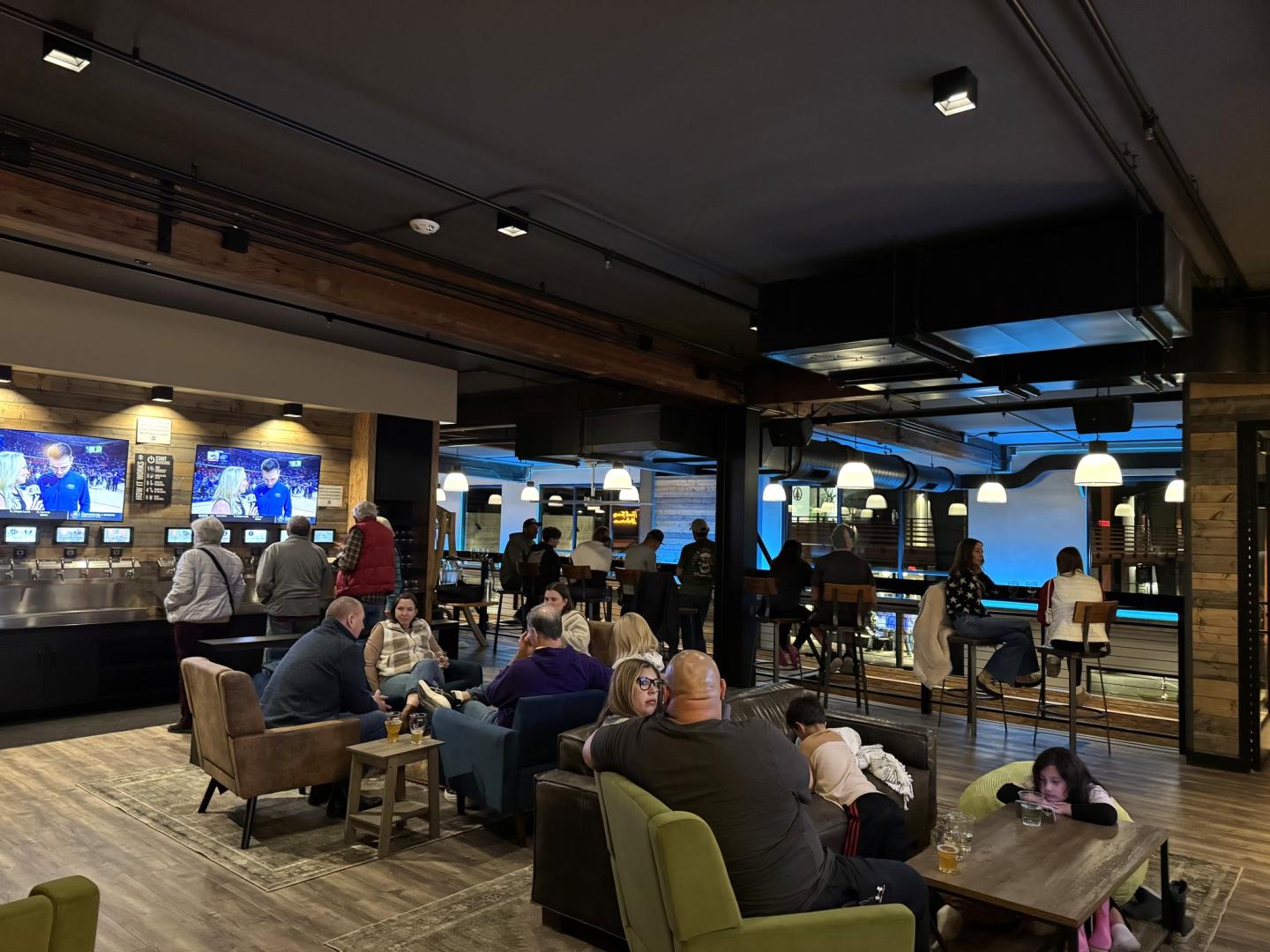 Cozy bar with people sitting and socializing, TVs on walls, warm lighting.