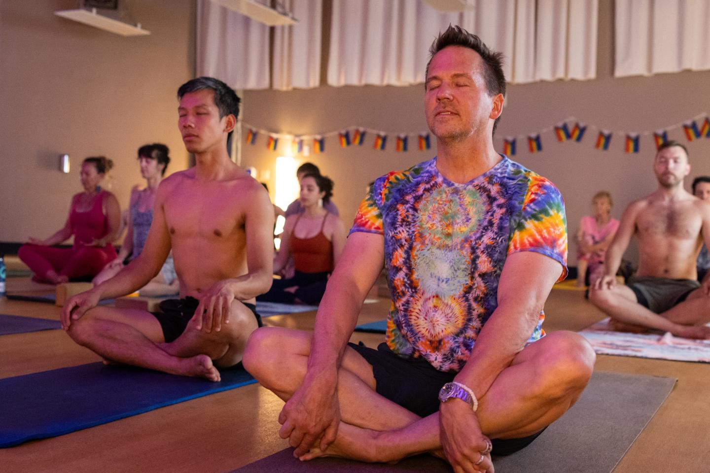 Group meditating in a studio, wearing colorful clothing, sitting cross-legged.