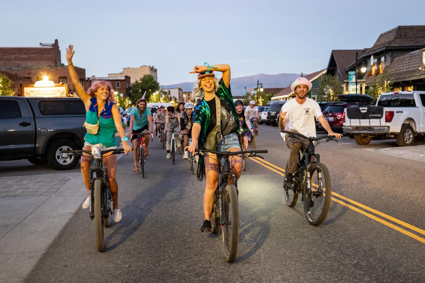 Cyclists ride down a street at dusk, some waving and smiling.