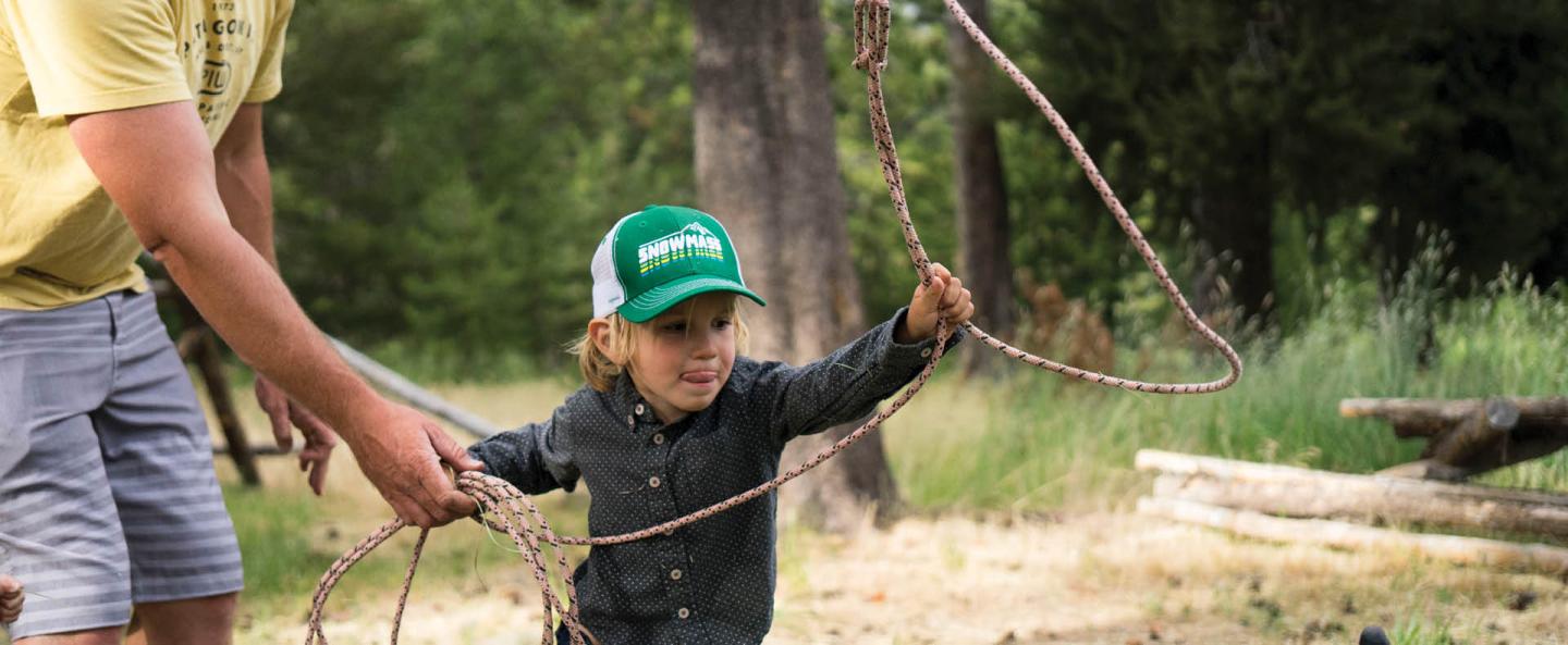 Child in green cap playing with lasso outdoors, assisted by an adult.