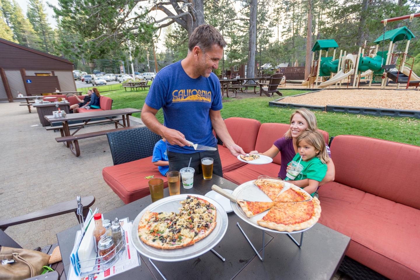 Man serving pizza to woman and child at outdoor seating area, park in the background.