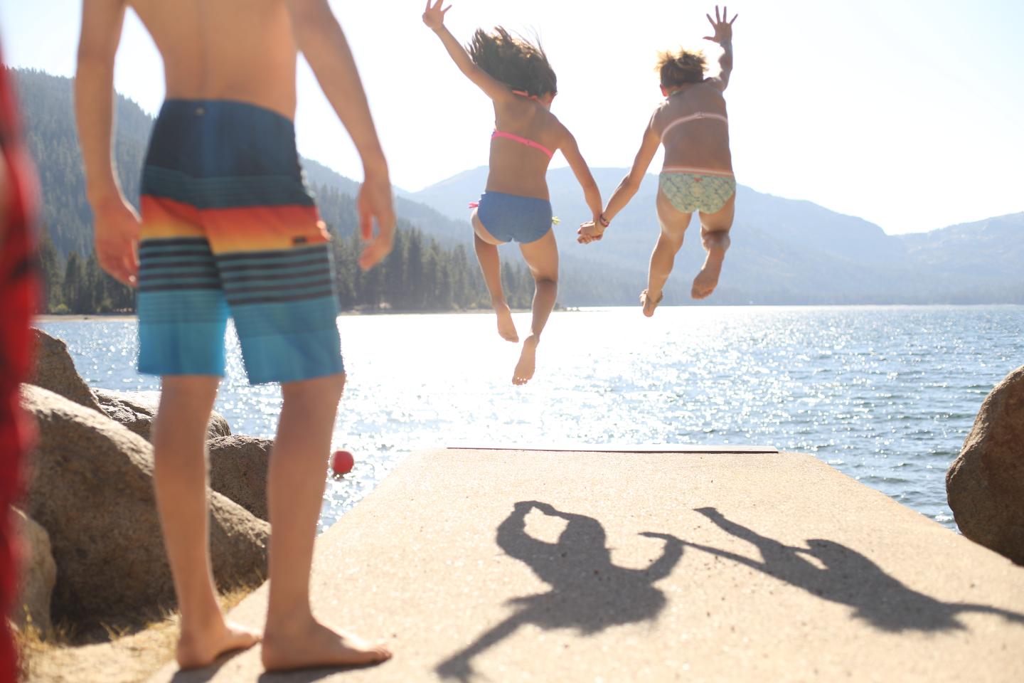 Kids jumping off a dock into a lake with mountains in the background.