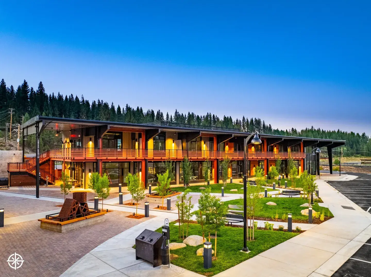 Modern lodge with wooden deck, lit warmly, surrounded by greenery at twilight.