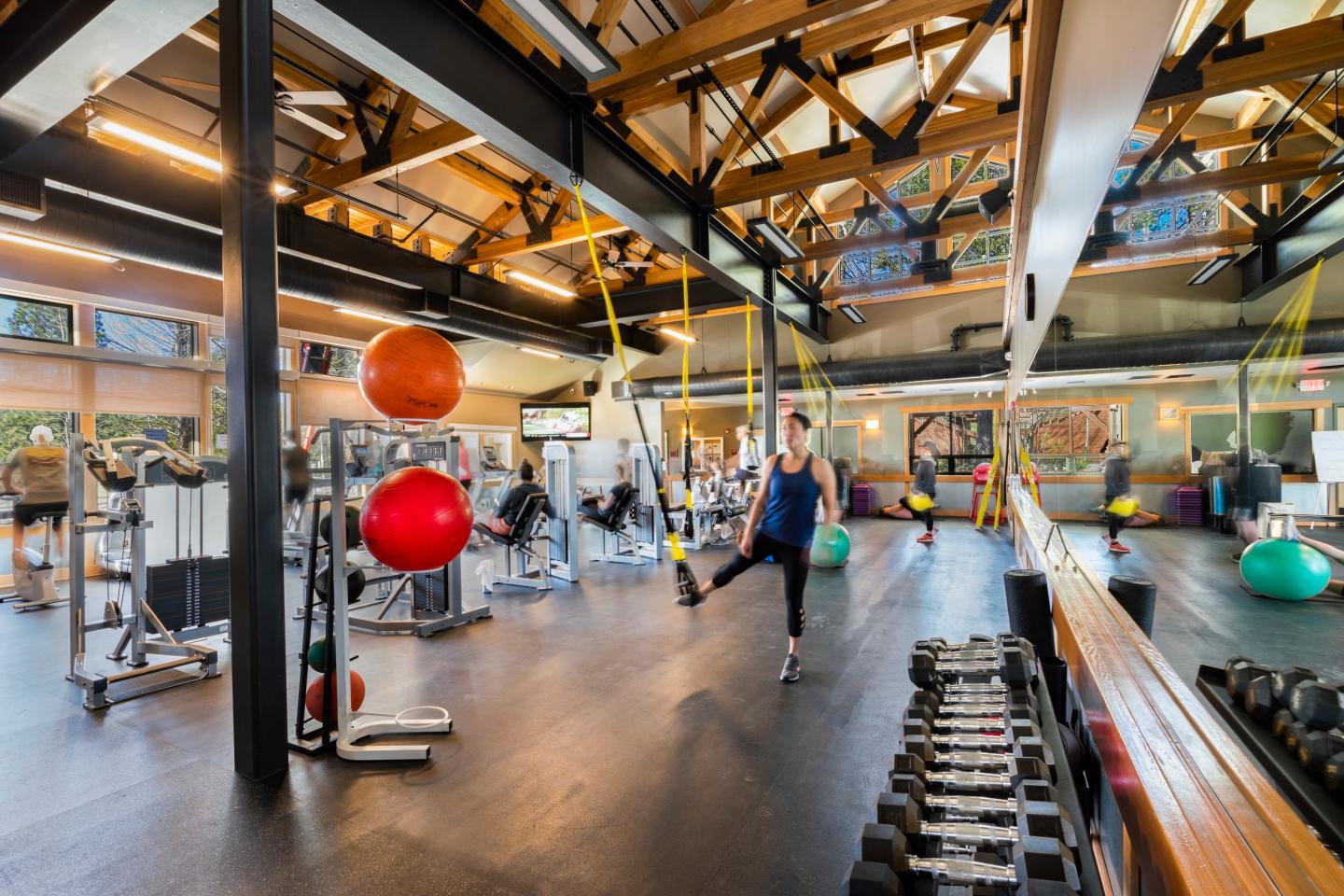 Spacious gym with people exercising, bright lighting, mirrors, and fitness equipment.