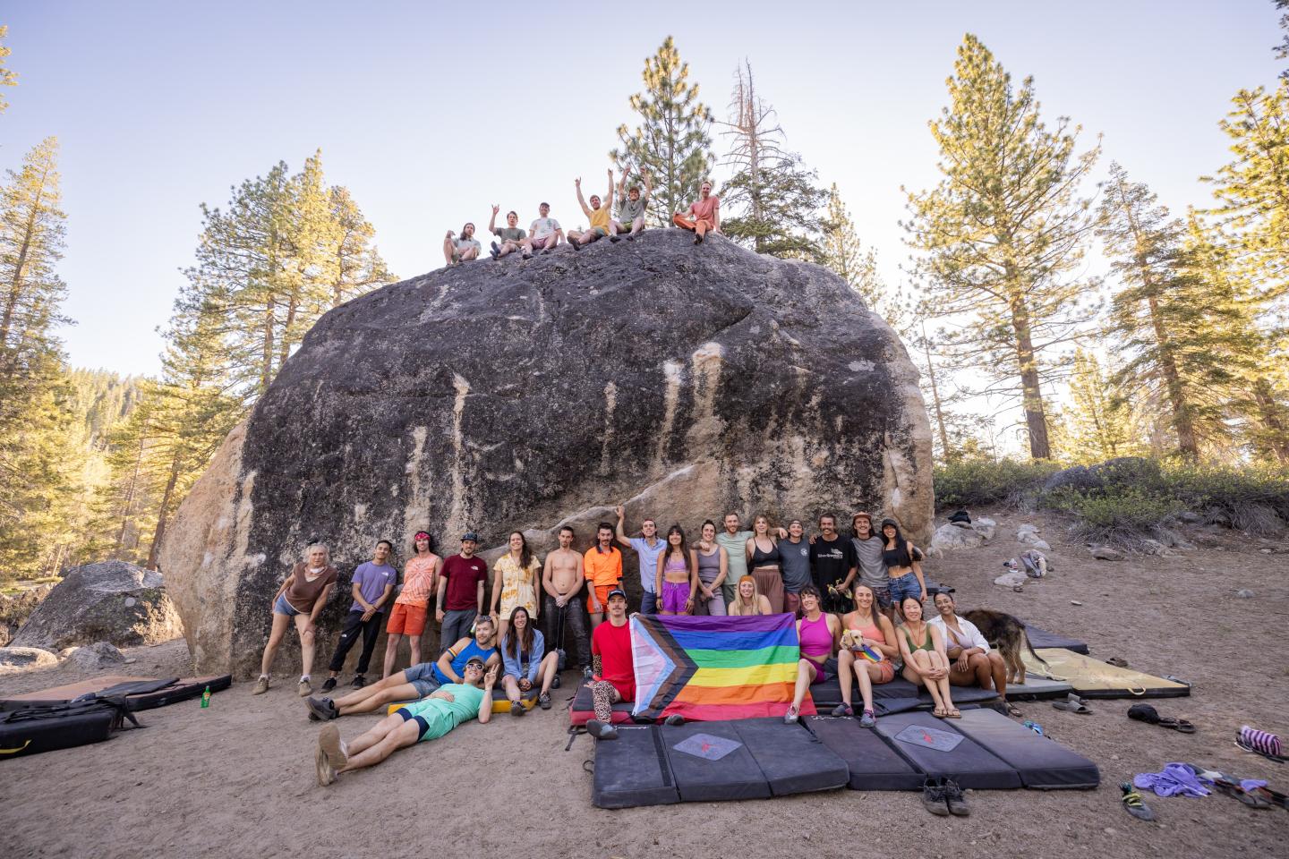Climbers gathered around and atop a large boulder in a forest setting, holding a pride flag.