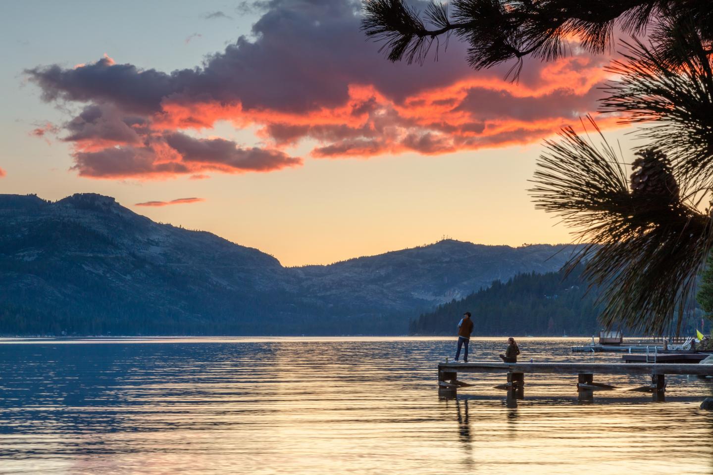 Sunset over a lake with mountains, dock, and silhouetted figures.