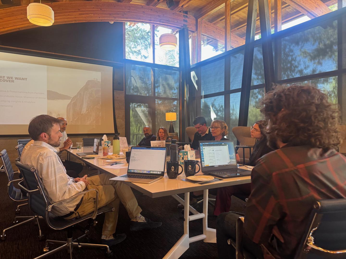 Meeting in modern room with large windows, people gathered around a table with laptops.