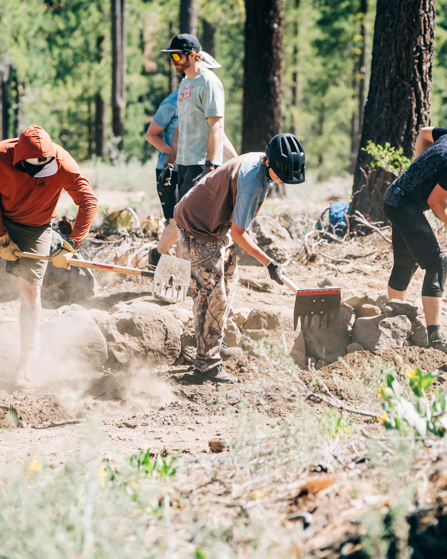 Cyclists digging dirt trail in forest, wearing helmets and casual clothes.