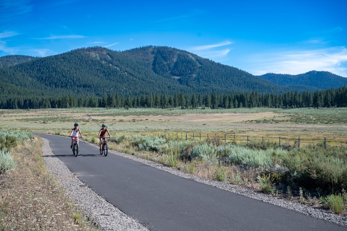 Two cyclists ride along a paved path amid grassy fields and mountains under a clear sky.
