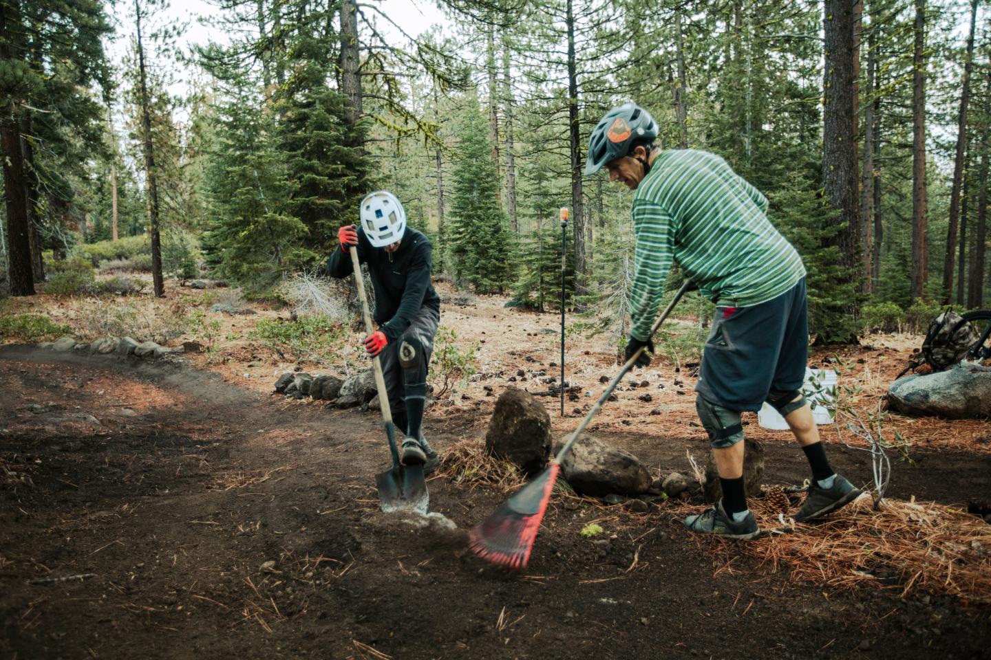 Two people in a forest, working on a dirt path with tools.