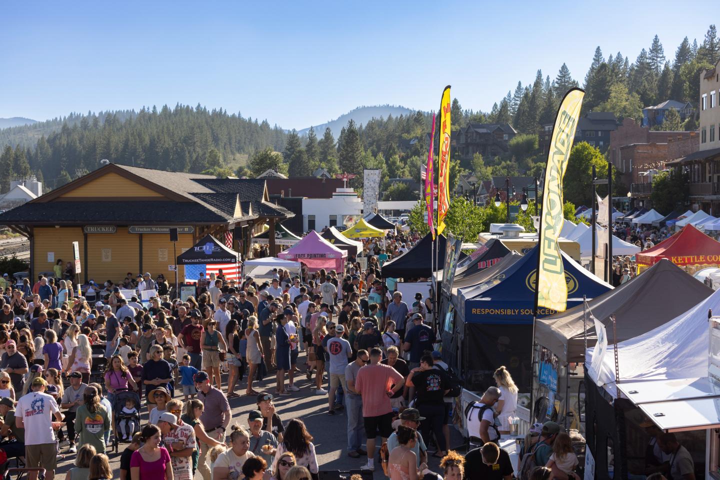 Crowd at an outdoor market with colorful tents and forested hills in the background.