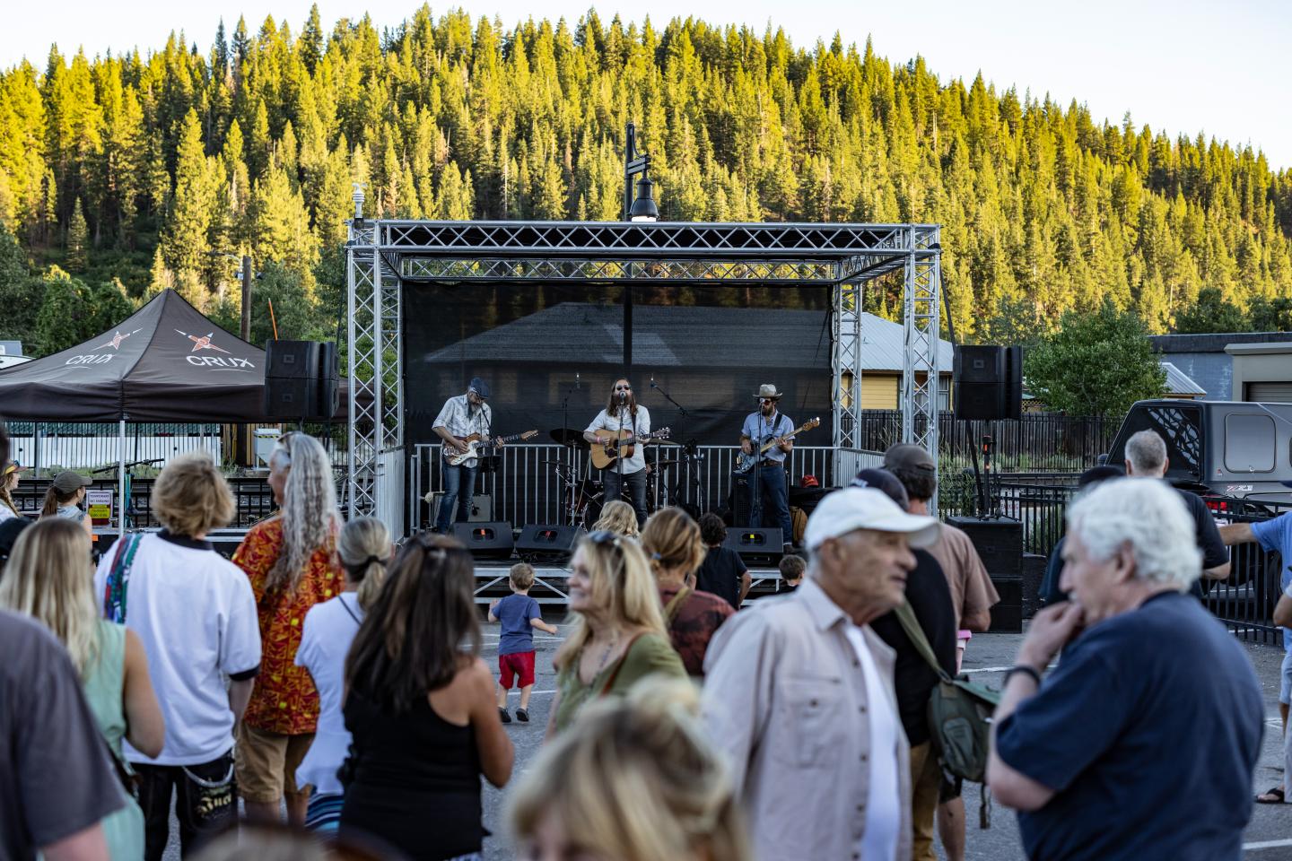 Outdoor concert with band on stage, crowd mingling, trees in the background.