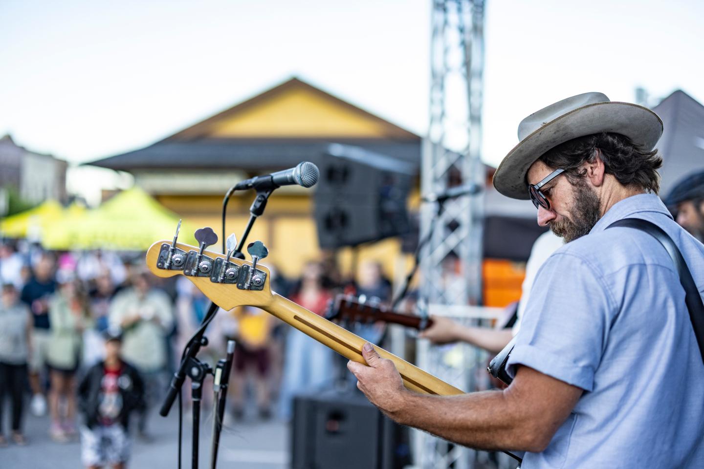 A musician plays guitar at an outdoor event, with a crowd in the background.
