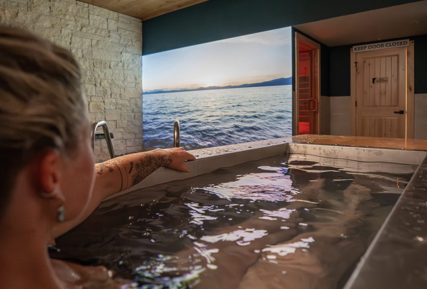 Person relaxing in a hot tub, pointing at a large ocean photo on the wall.