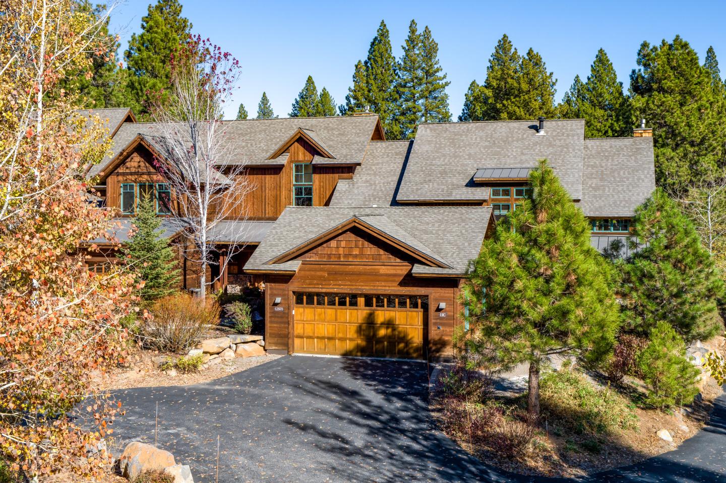 Rustic wooden house with a shingled roof, surrounded by trees and a driveway.