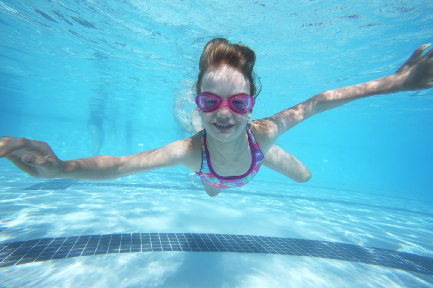 Child swimming underwater in a pool, wearing pink goggles and swimsuit.