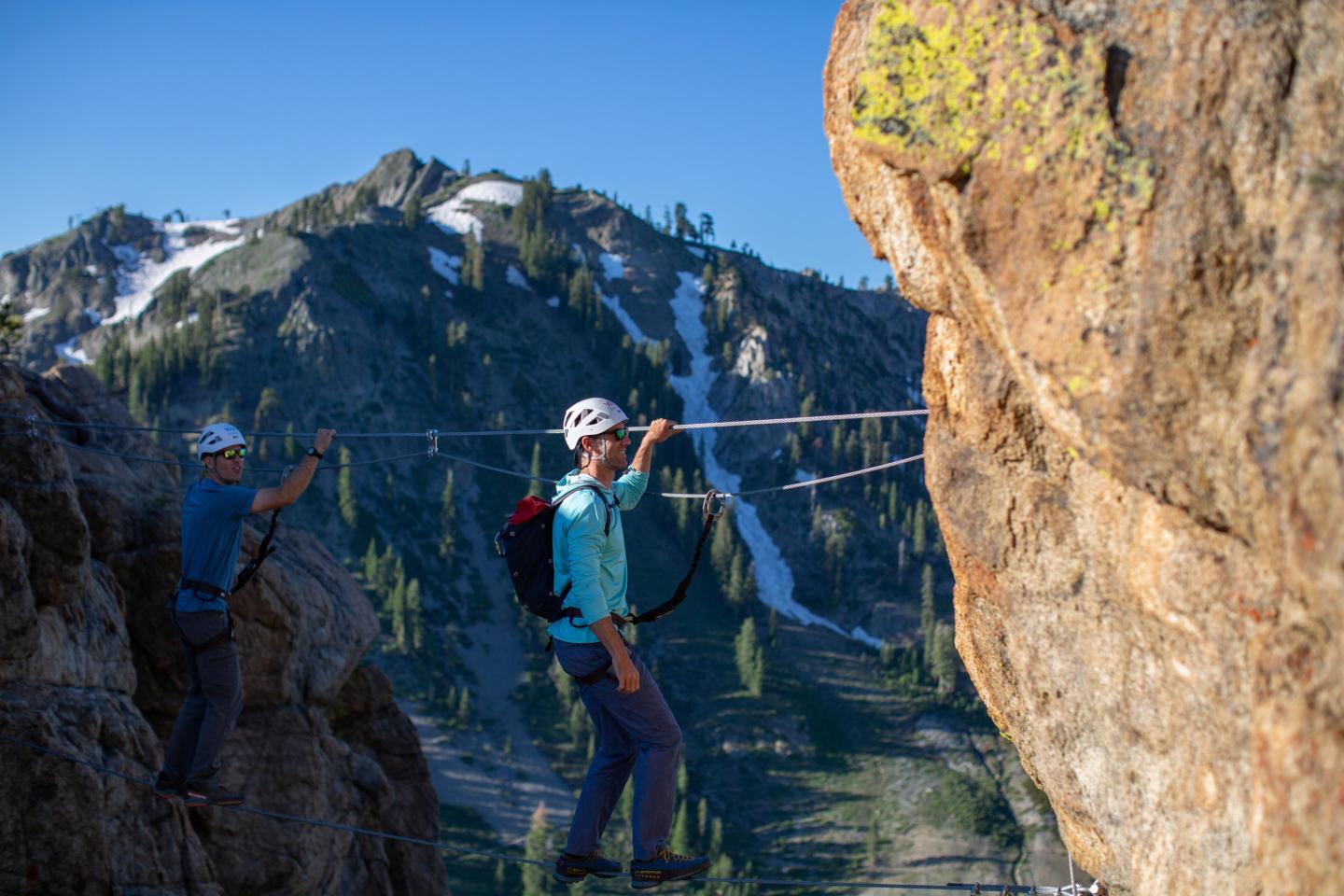 Two people in helmets crossing a rope bridge between rocky cliffs, with mountains in the background.