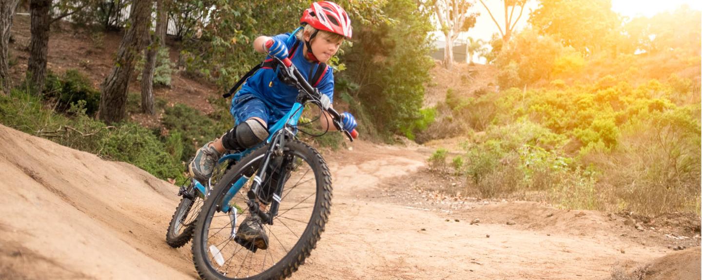 Child mountain biking on a dirt trail in the forest, sunlight filtering through trees.