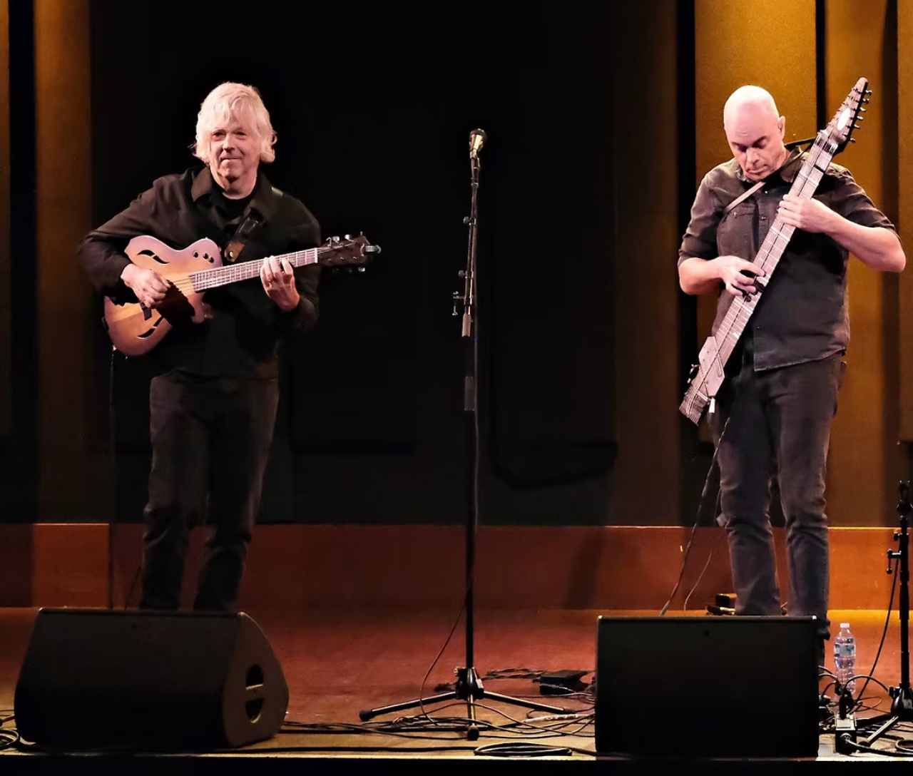 Two musicians perform on stage, one with a guitar, the other with a bass.