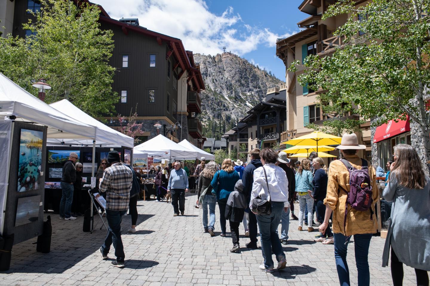 Outdoor market with people walking, tents, and mountain backdrop on a sunny day.
