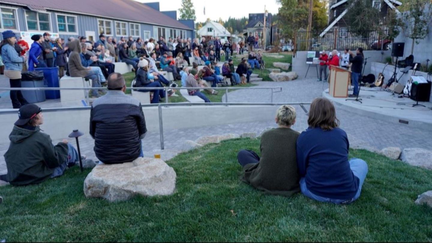 Outdoor gathering with seated crowd, speakers at a podium, evening setting.