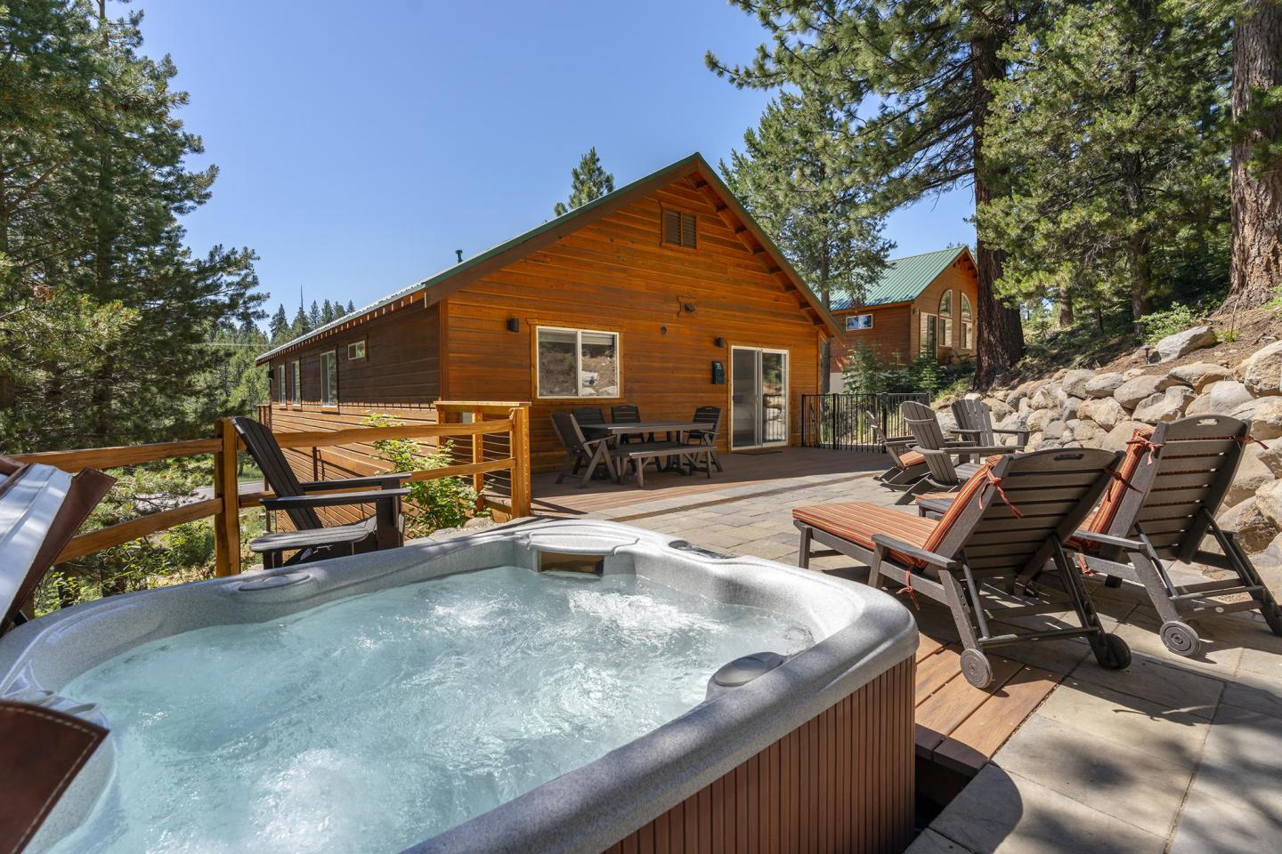 Hot tub on a wooden deck with chairs, near a cabin, surrounded by trees.