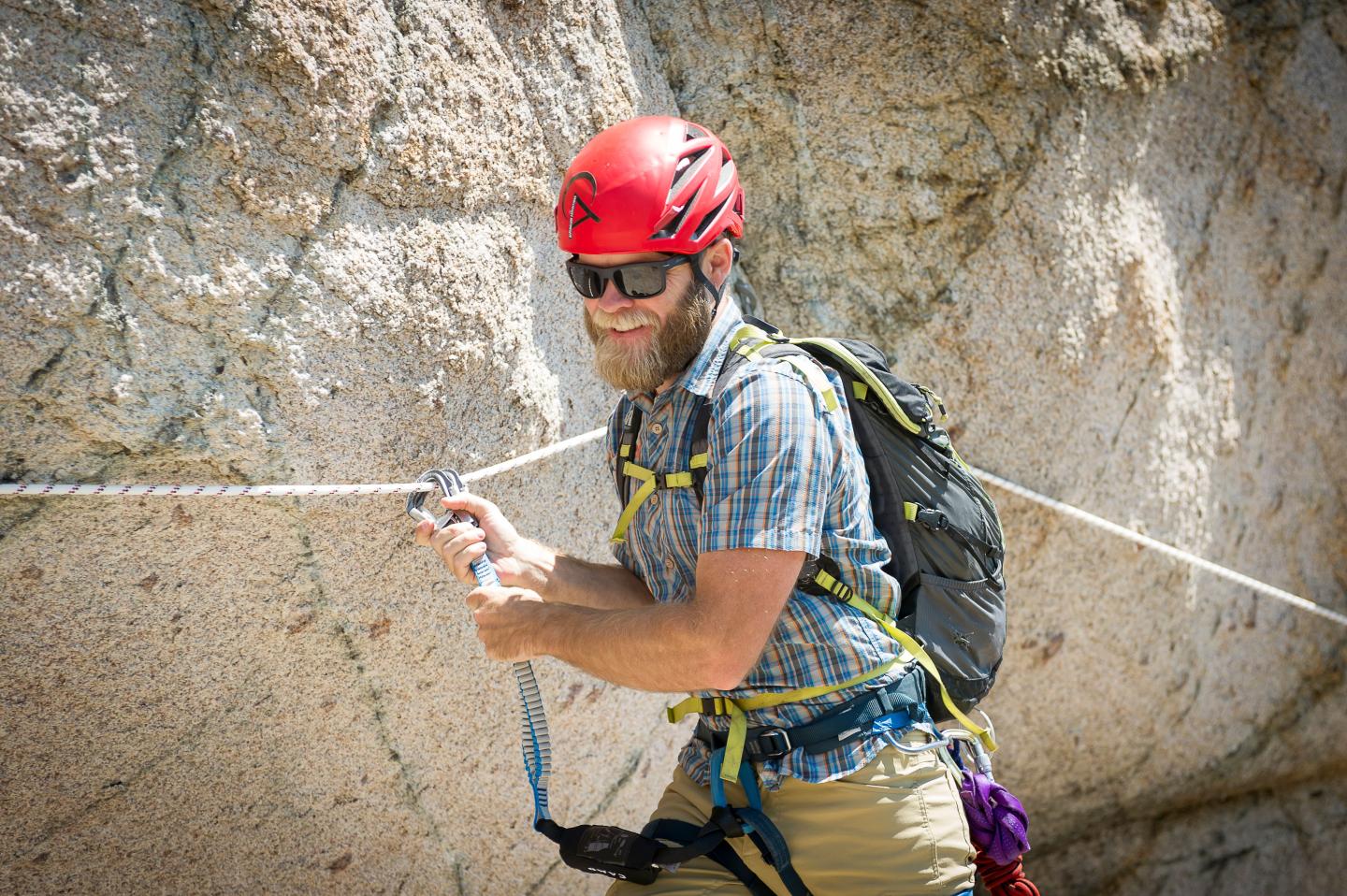 Rock climber with a red helmet smiling, standing on a ledge.