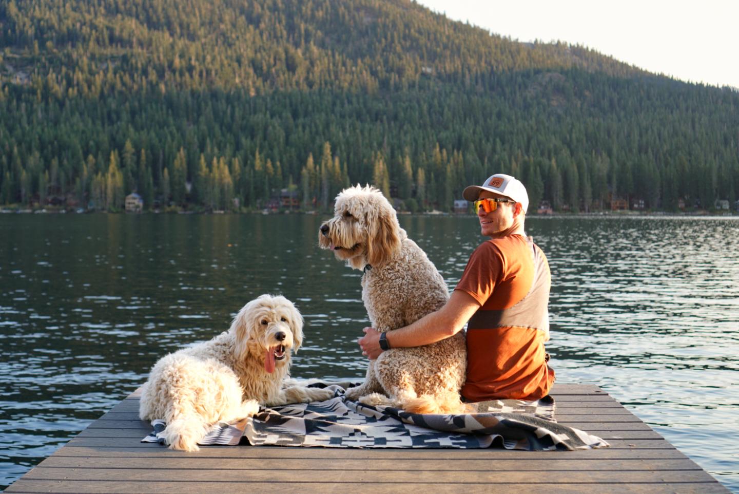 Man with two dogs sitting on a lakeside dock, forested hills in the background.