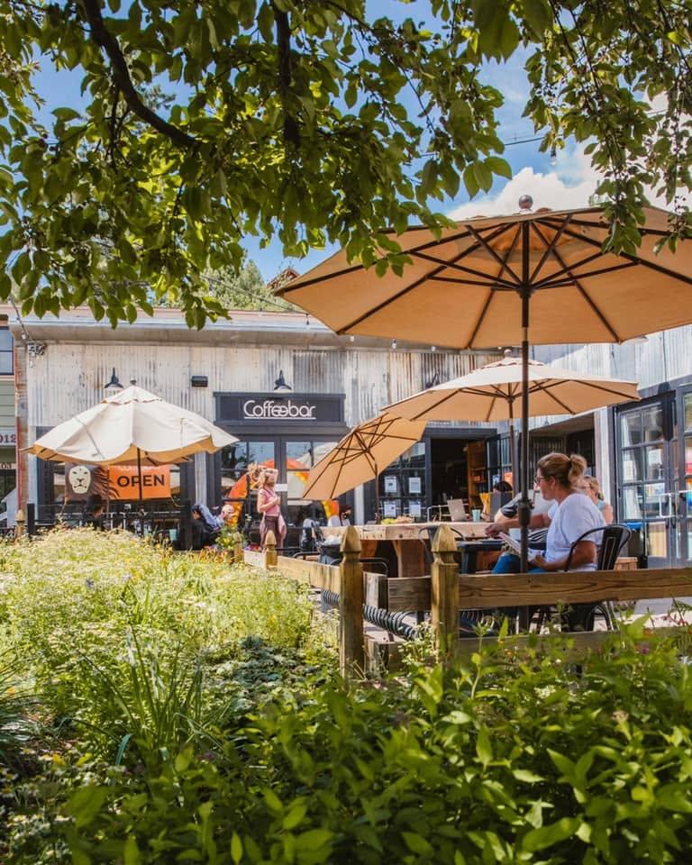 Outdoor café with large umbrellas and people dining, surrounded by greenery.