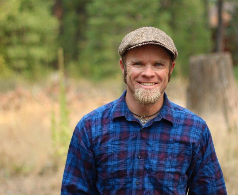 Man with a beard in a blue plaid shirt and cap, standing in a forest.