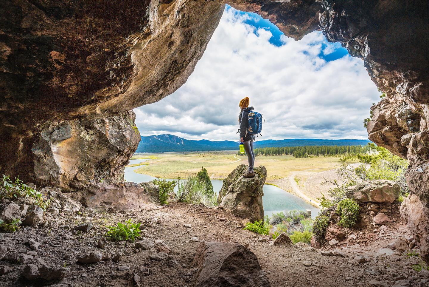 Hiker standing in a cave opening, overlooking a scenic valley and river.