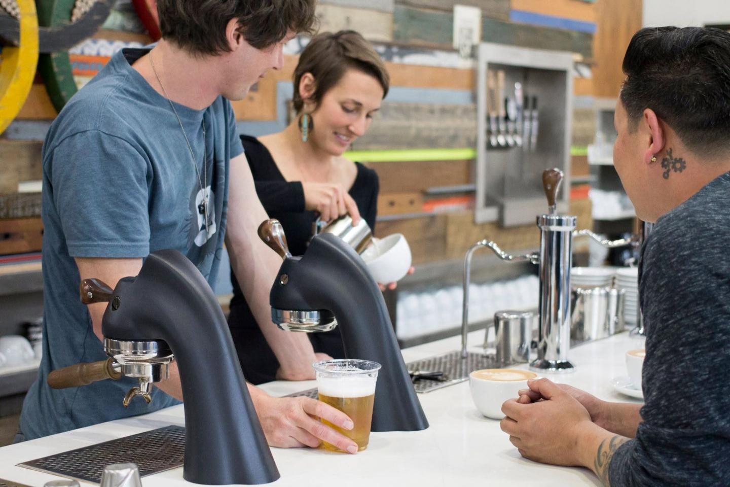 Barista pours milk into coffee, two people watch, modern cafe setting.