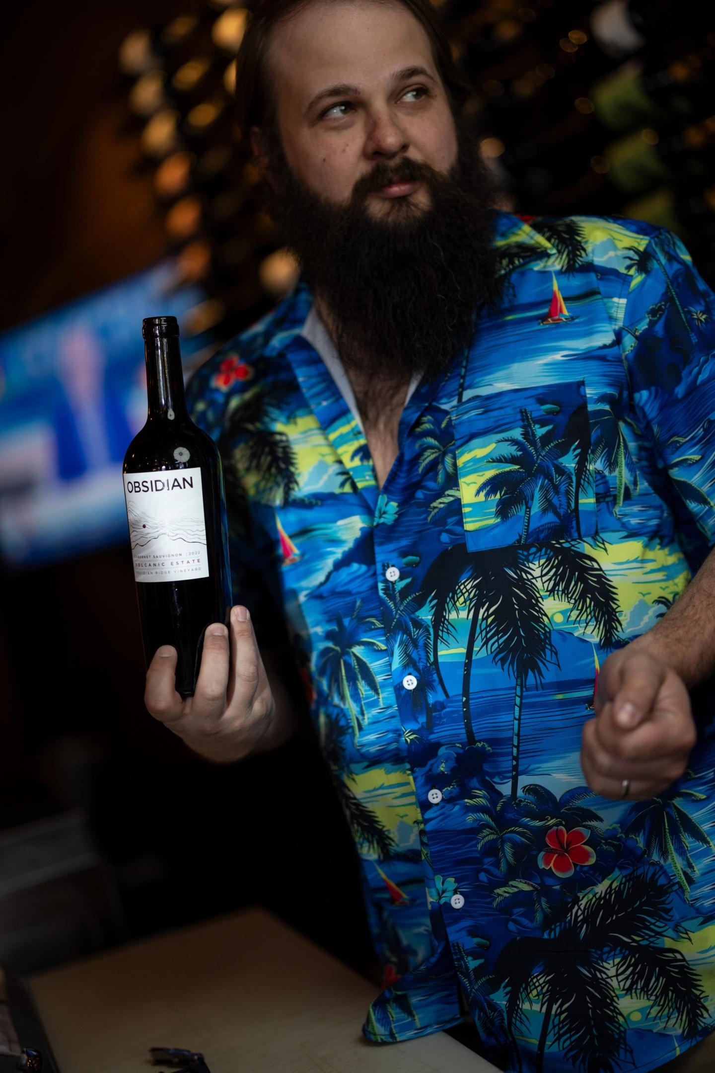 Man in a tropical shirt holding a wine bottle indoors.