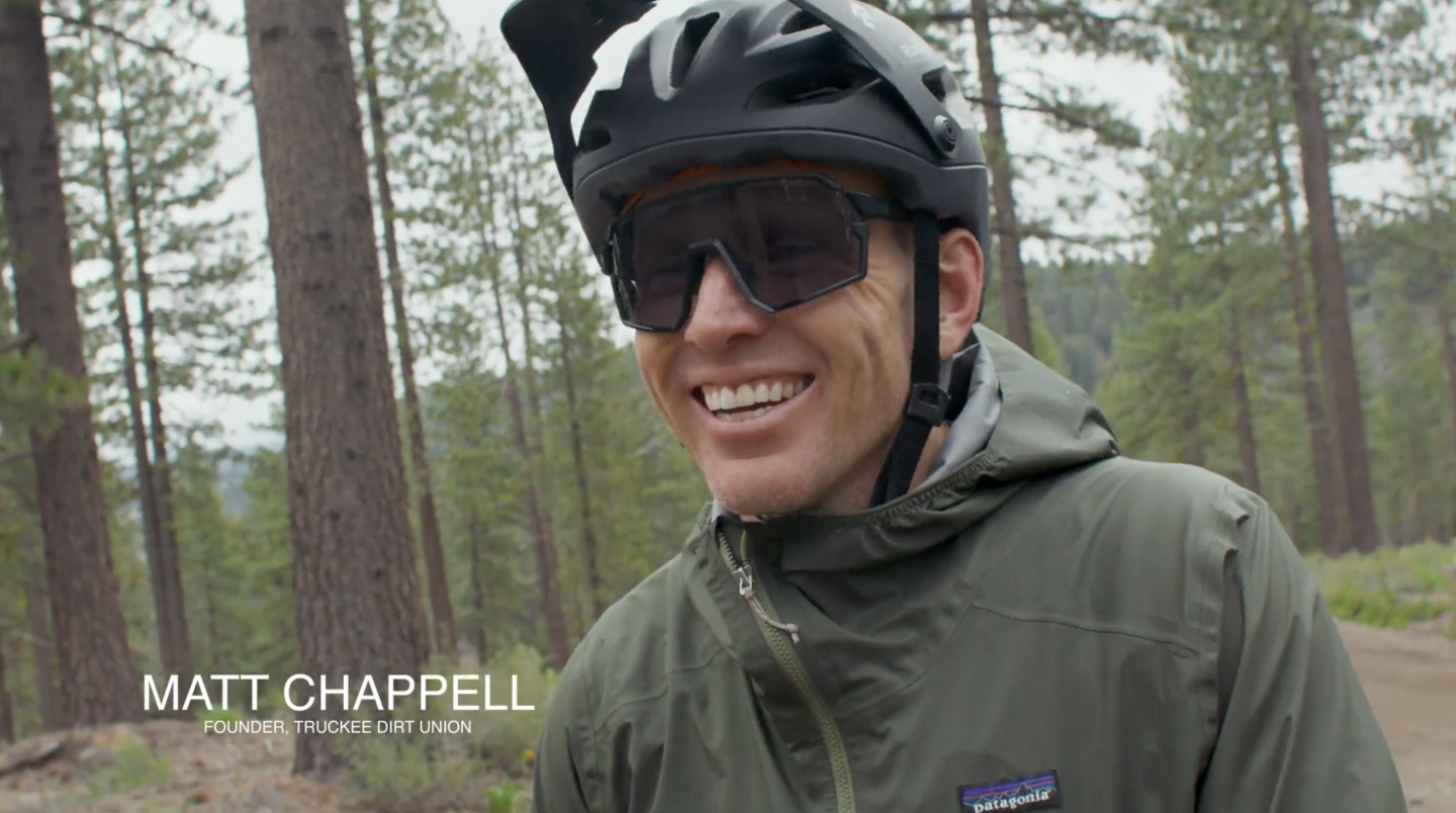 Smiling person in a bike helmet and sunglasses in a forest setting.