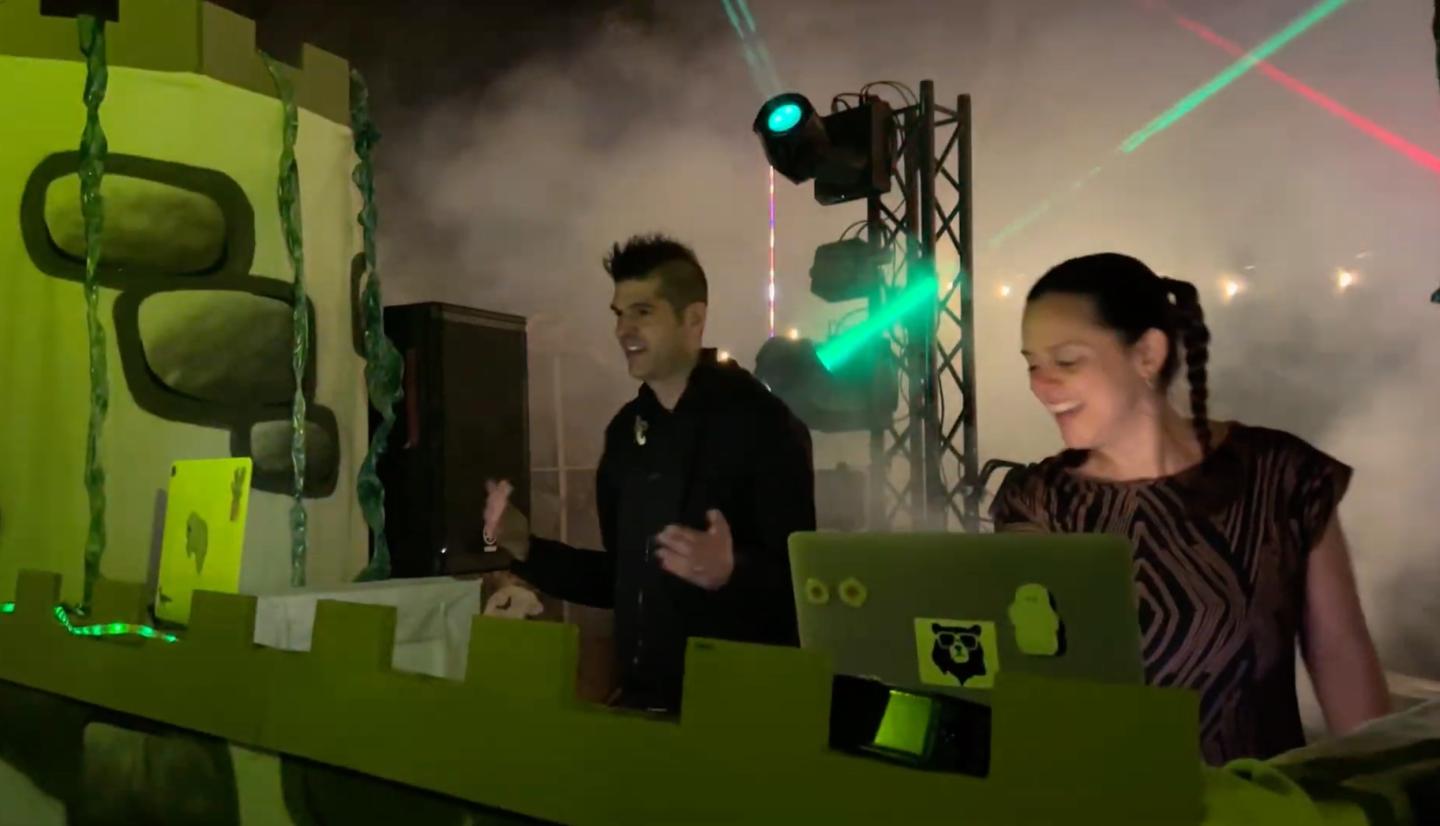 DJ duo playing music in a green-lit, smoke-filled club with colorful lights.