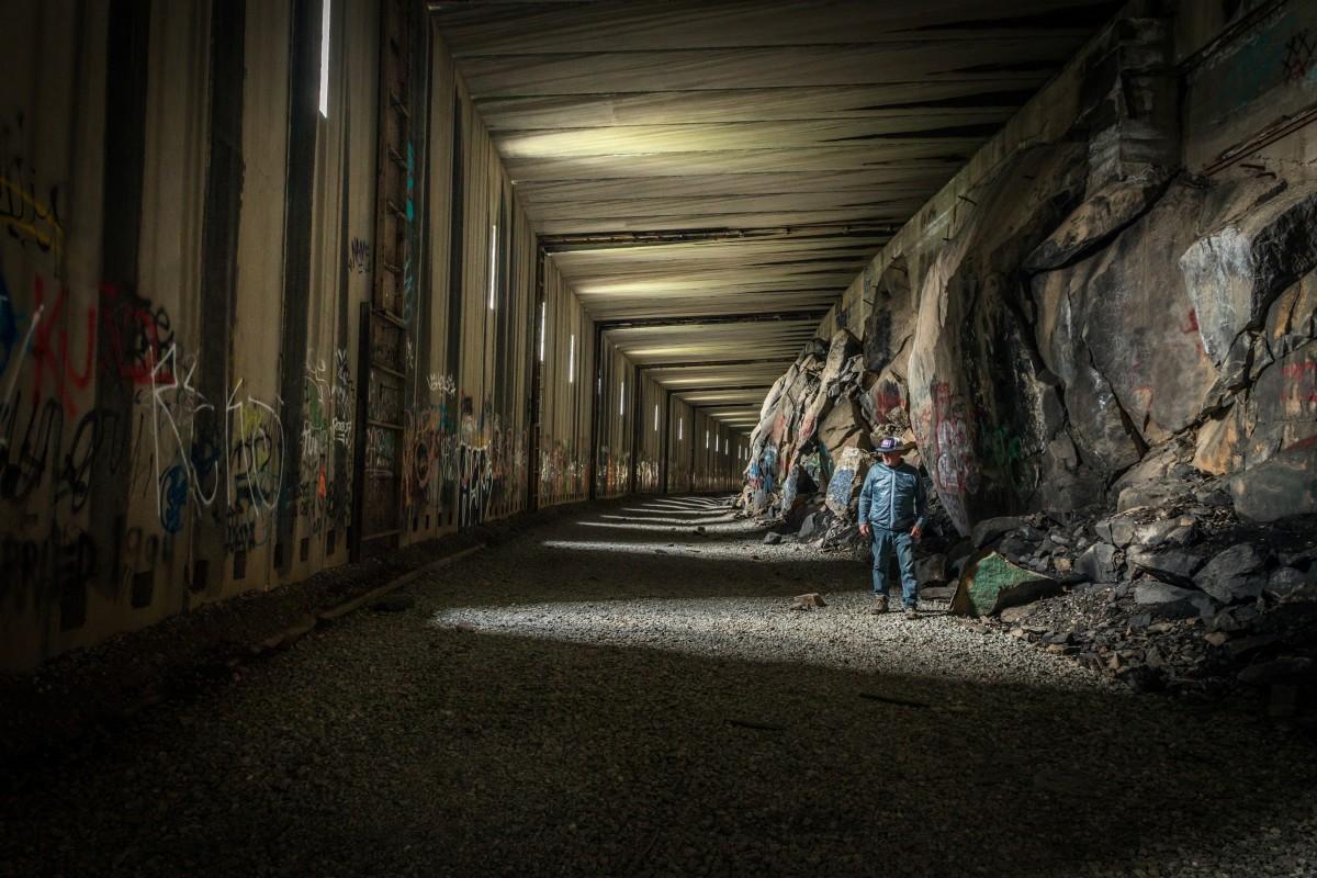 Dark tunnel with graffiti, rocky walls, and two people walking. Light filters from above.