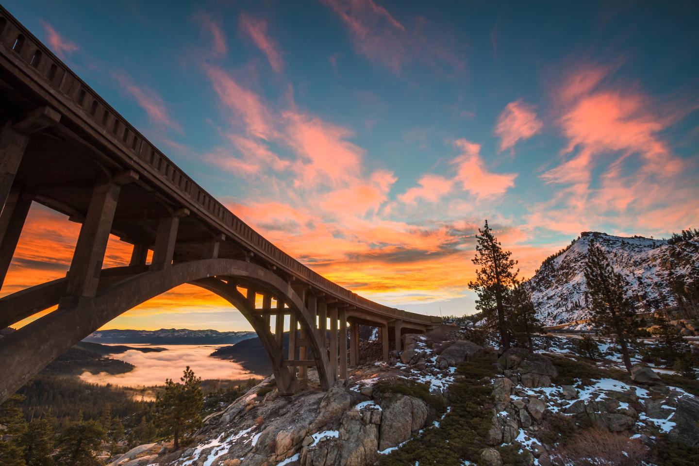 Bridge at sunset with colorful sky and snowy mountains.