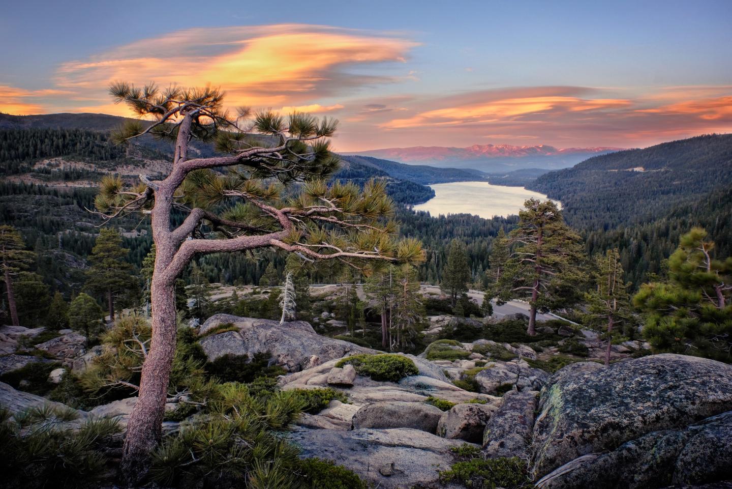 Mountain landscape at sunset with trees, rocks, and a distant lake.
