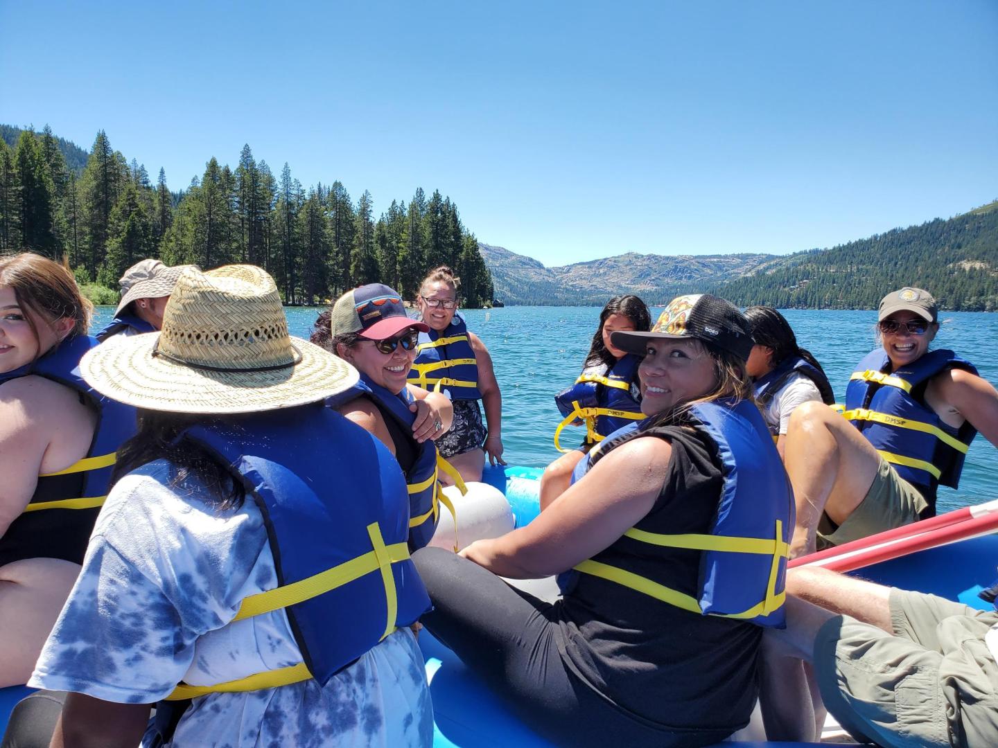 People wearing life vests sitting in a raft on a lake, surrounded by mountains and trees.