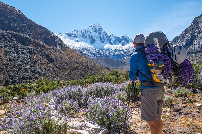 Hiker with backpack facing snow-capped mountain under blue sky.