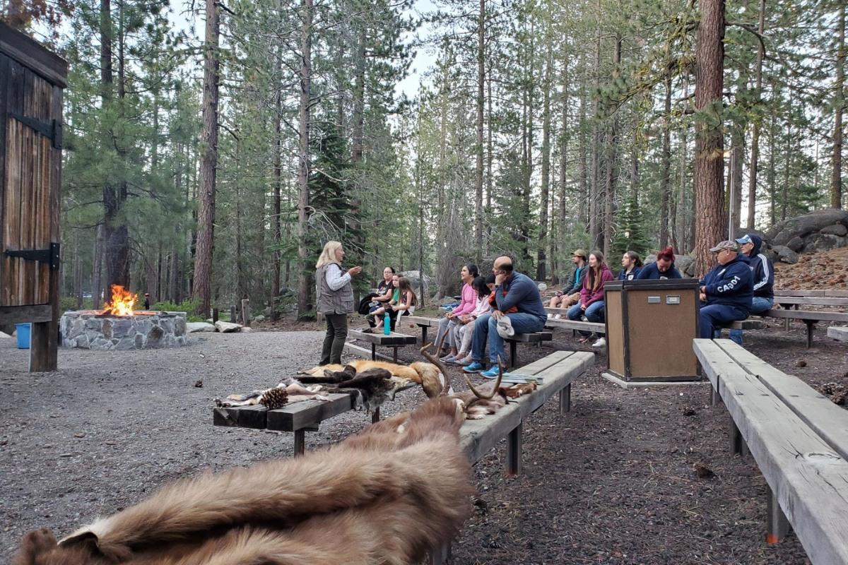 Outdoor campfire talk in a forest, group seated on benches facing speaker.
