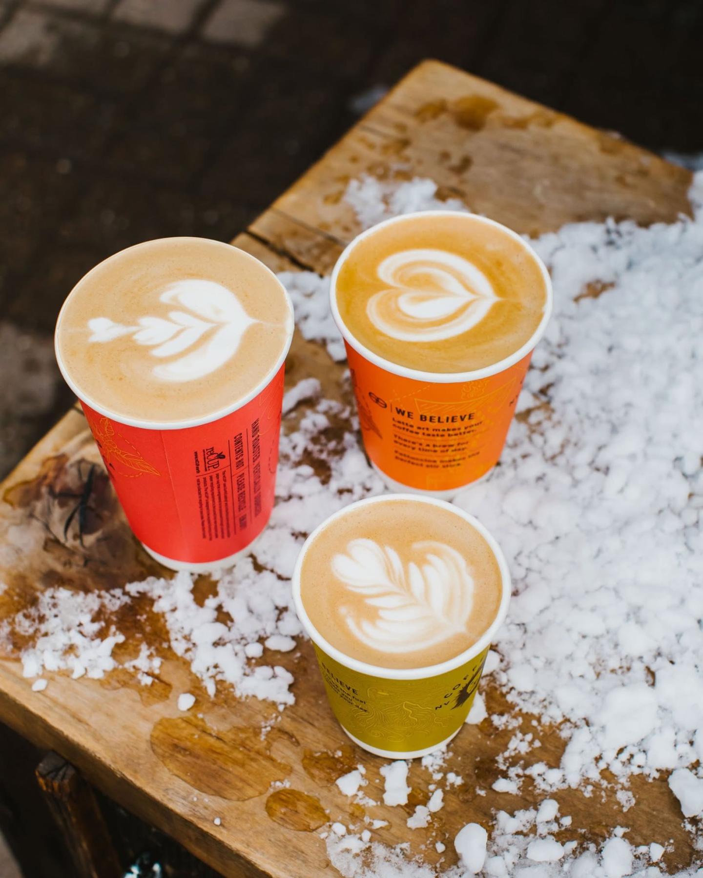 Three cups of latte with heart latte art on a snowy table outside.